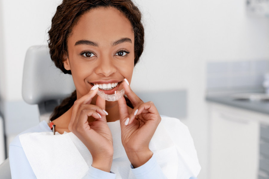 Pretty black girl at the dental office holding a clear aligner in front of her smile.